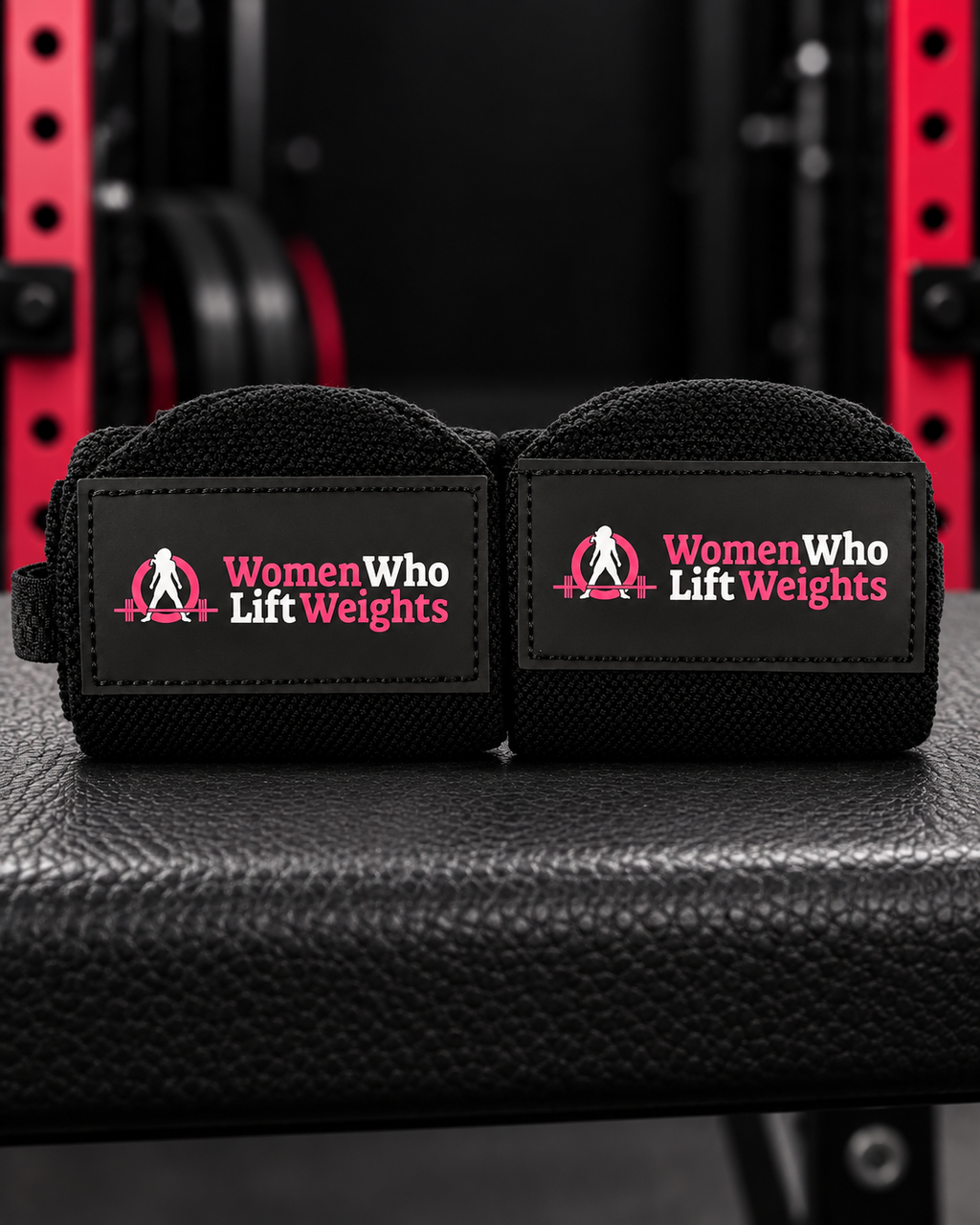 Pair of black Women Who Lift Weights wrist wraps with pink and white logo patches.